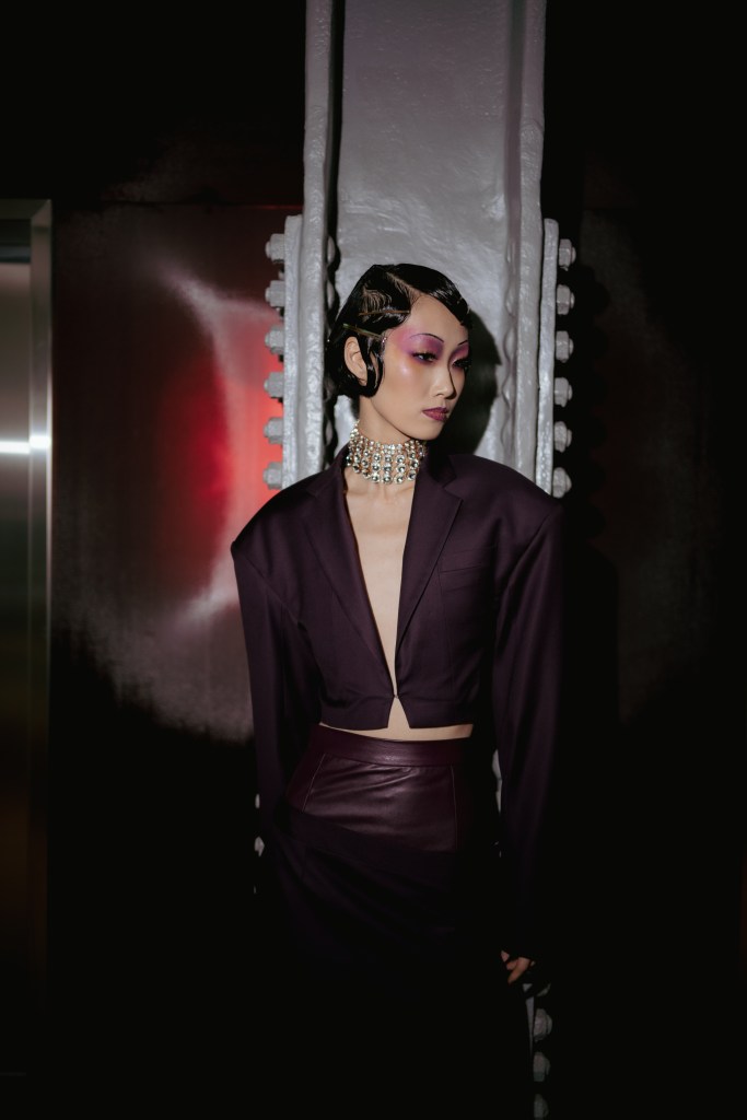 Shao New York Fall/Winter 2025 runway look featuring a cropped deep purple blazer with structured shoulders and a matching high-waisted leather-paneled midi skirt. The bold ensemble is accented by dramatic purple makeup, a sparkling statement choker, and sleek, sculpted hair. This look embodies a fusion of futuristic elegance and power dressing, highlighting the brand’s avant-garde approach to tailoring.