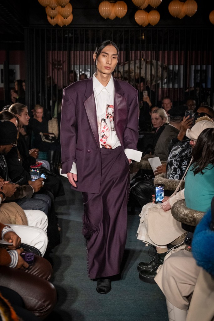 Shao New York Fall/Winter 2025 runway look featuring an oversized, deep purple blazer and matching wide-leg trousers. The ensemble is paired with a statement white shirt adorned with a bold graphic print. The look exemplifies modern tailoring with a contemporary, artistic twist, showcasing the brand's signature fusion of avant-garde silhouettes and streetwear influences.